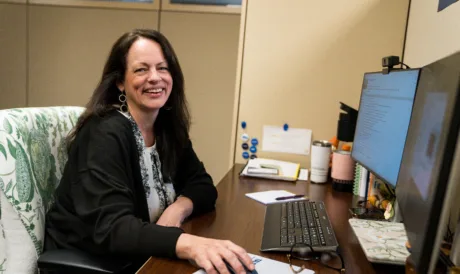 Amy Metz, smiling at the camera while browsing on the computer at her desk.